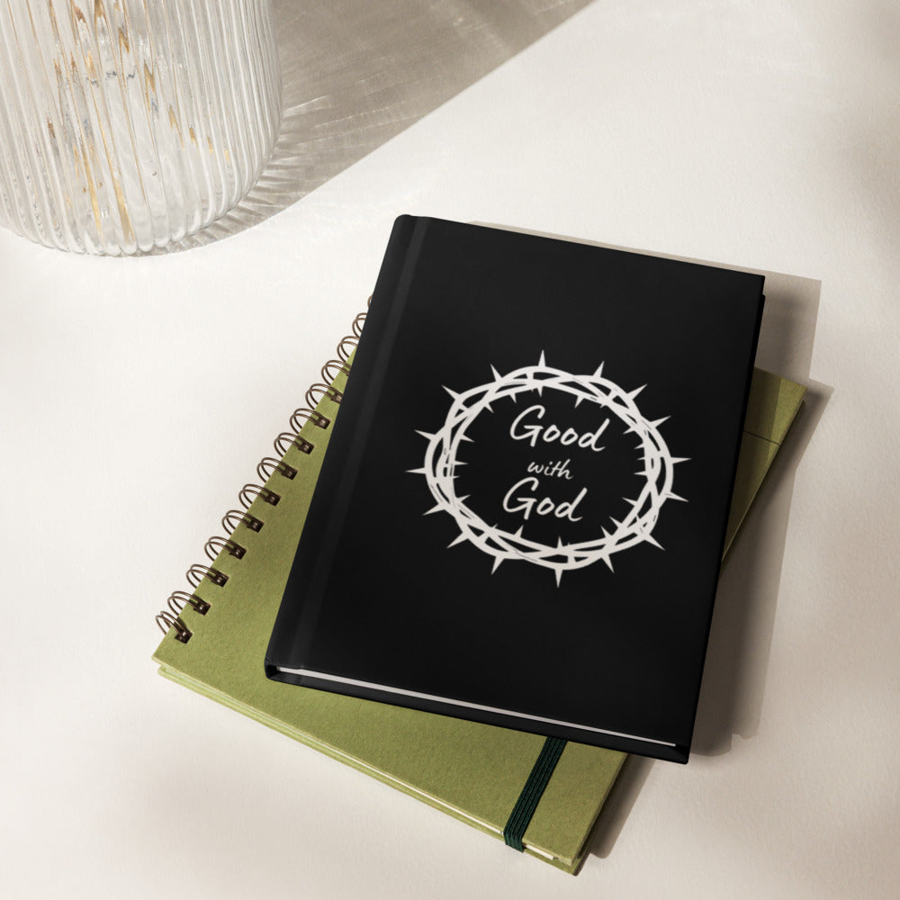 A black hardcover notebook with a silver-toned ornate crest and the words, "Good with God" on the cover, resting on top of a light olive-green spiral notebook on a white surface.