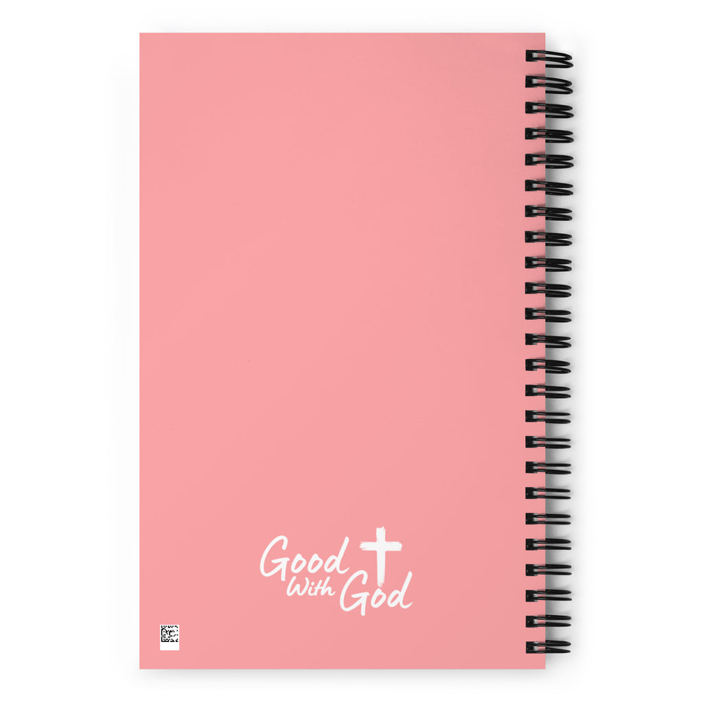 A pink spiral notebook with a black wire coil binding and the words 'Good with God' printed in white near the bottom, alongside a white cross graphic.